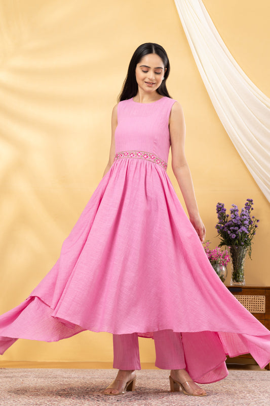 A woman wearing a pink floral embroidered kurta set with a asymmetrical hemline, paired with straight pants. She is also wearing floral earrings, beige heels, and carrying a handbag.